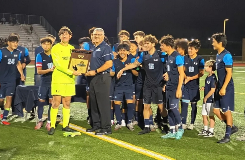 A soccer team celebrates on a field at night, holding a championship trophy.
