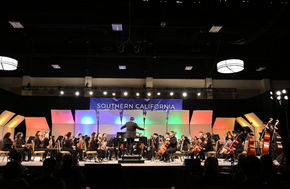 A conductor leads a large orchestra on a stage with colorful lighting and a banner reading 'SOUTHERN CALIFORNIA SCHOOL BAND AND ORCHESTRA ASSOCIATION'.