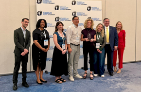 A group of eight people stand together in front of a banner for the "Student and Community Voice AI Summit."
