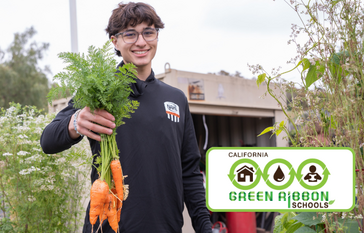 A smiling student holds a bunch of freshly harvested carrots, showcasing produce from a school garden.
