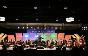A conductor leads a large orchestra on a stage with colorful lighting and a banner reading 'SOUTHERN CALIFORNIA SCHOOL BAND AND ORCHESTRA ASSOCIATION'.