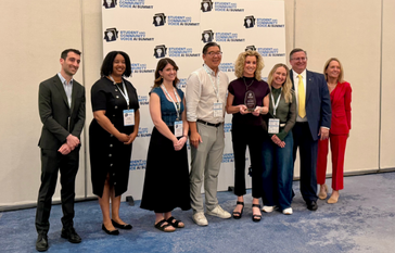 A group of eight people stand together in front of a banner for the "Student and Community Voice AI Summit."