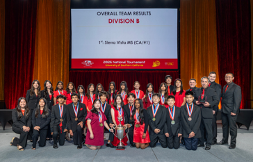 A group of students and adults in formal attire pose for a photo on stage, celebrating a first-place victory at the 2026 National Tournament.