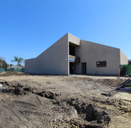 A ground-level view of a construction area showing dug-out earth and utility markings leading toward the angular entrance of a school building.