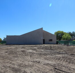 A wide shot of a cleared, dirt construction site in front of a tan, modern school building with a distinct sloped roofline under a clear blue sky.
