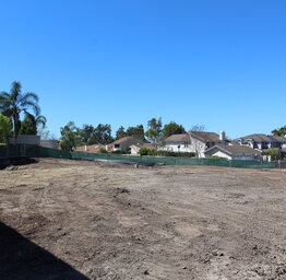 A wide shot of a cleared, dirt construction site enclosed by green mesh fencing with suburban houses and palm trees in the background under a clear blue sky.