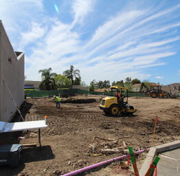 Construction workers and a yellow steamroller level a dirt lot next to a performing arts building under a streaky blue sky.