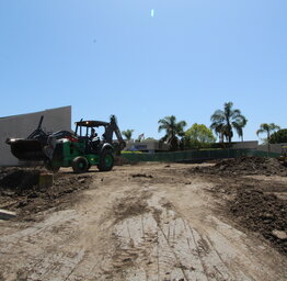 A green tractor with a backhoe attachment working at a construction site in front of school buildings.