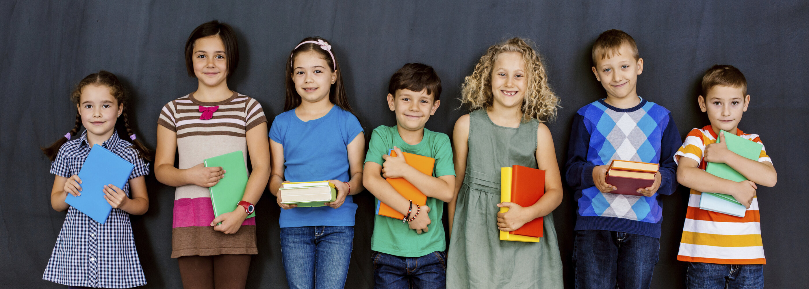 Seven smiling children of various heights stand in a line against a dark wall, each holding a book