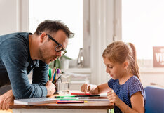 A man with glasses leans over a wooden desk to help a young girl with her schoolwork in a sunlit room