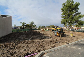 A John Deere track loader sits on a muddy construction site near a large pile of dirt and green fencing.