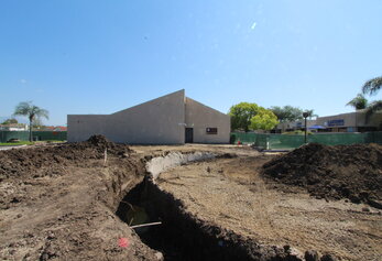 Earthwork progresses at a school construction site, featuring excavated trenches and large mounds of dark soil near a modern sloped building.