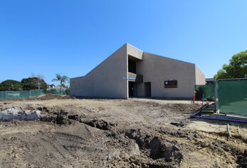 A ground-level view of a construction area showing dug-out earth and utility markings leading toward the angular entrance of a school building.