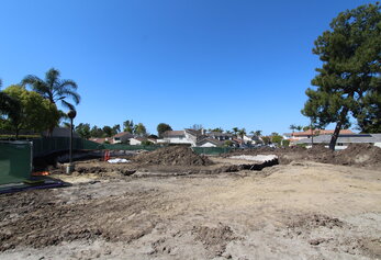 An active construction site features a curved excavation trench and piles of dark soil against a backdrop of a green safety fence and a residential neighborhood.