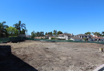 A perspective view of a large, leveled dirt field surrounded by green mesh fencing and a row of suburban two-story homes.