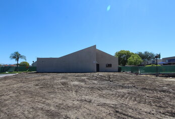 A wide shot of a cleared, dirt construction site in front of a tan, modern school building with a distinct sloped roofline under a clear blue sky.