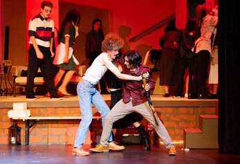 Students perform a tense scene from Portola High School’s Night of the Living Dead, with one student restraining another under red lighting.
