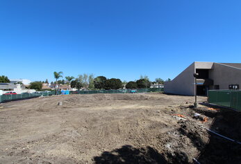 A view of a construction area featuring excavated dirt mounds, blue portable toilets in the distance, and the side of a modern school building under a bright sun.