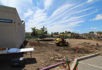 Construction workers and a yellow steamroller level a dirt lot next to a performing arts building under a streaky blue sky.