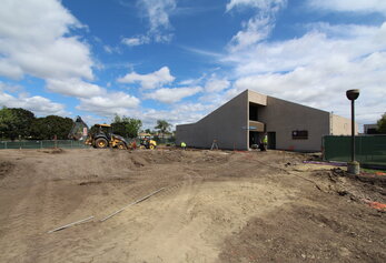 A yellow backhoe and a smaller loader work on an expansive dirt excavation site in front of a modern school building.