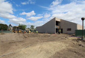 Heavy machinery operates on a large dirt construction site with several workers near the entrance of an angular tan building.
