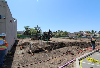 Two construction workers and a green backhoe on a dirt lot, with the Lakeside Middle School building in the background.