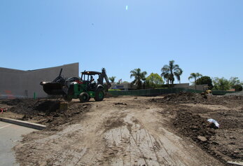 A green tractor with a backhoe attachment working at a construction site in front of school buildings.