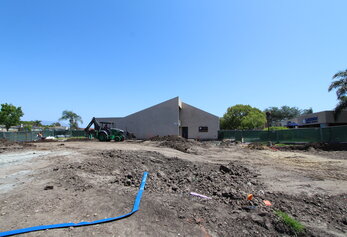 A wide view of a construction site featuring piles of dirt, a green backhoe, and Lakeside Middle School in the background.