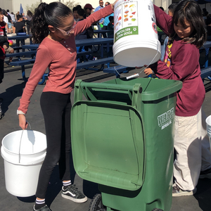 Two girl students dumping buckets of food waste into a bigger green trash can