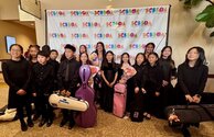 A group of young musicians in black attire pose with their instruments and bouquets in front of a SESBOA banner.