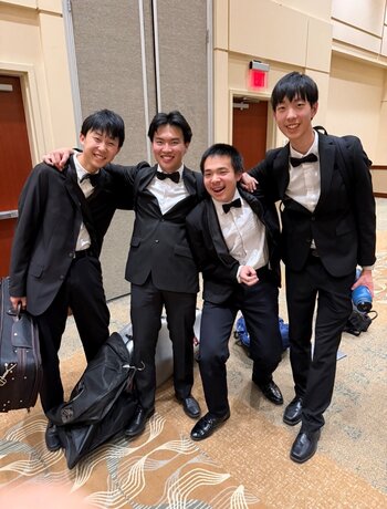 Four young men in matching black suits and bow ties pose for a photo, smiling.