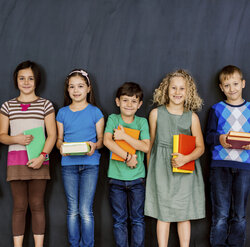 Seven smiling children of various heights stand in a line against a dark wall, each holding a book